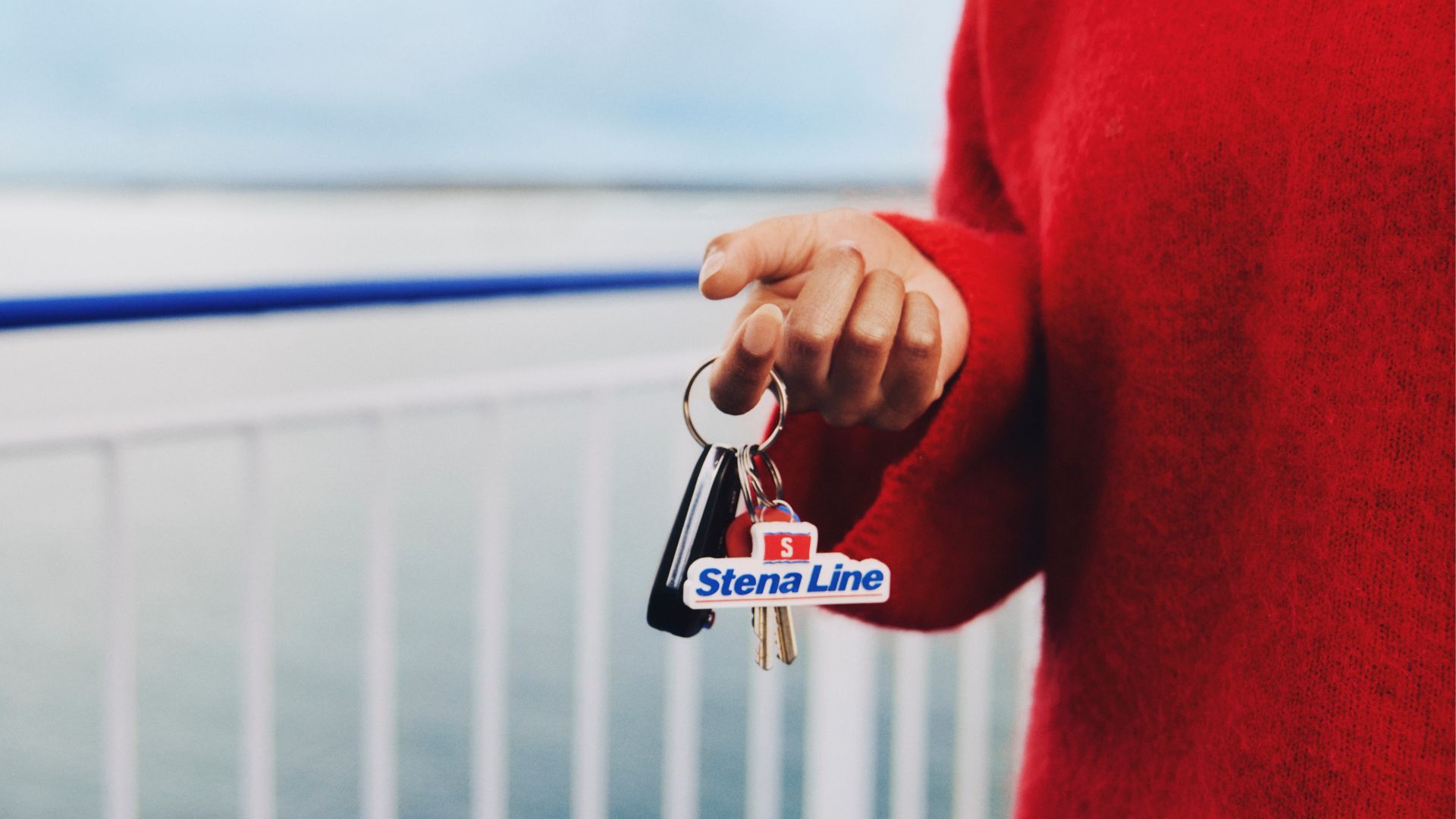 A person in a red sweater holds a set of keys with a clearly visible Stena Line keychain. The scene is set on an outdoor ferry deck with a calm sea and railing in the background. The image conveys a relaxed, travel-oriented mood with cool blue and red tones. The focus is on the hand and keychain, with no other people present.
